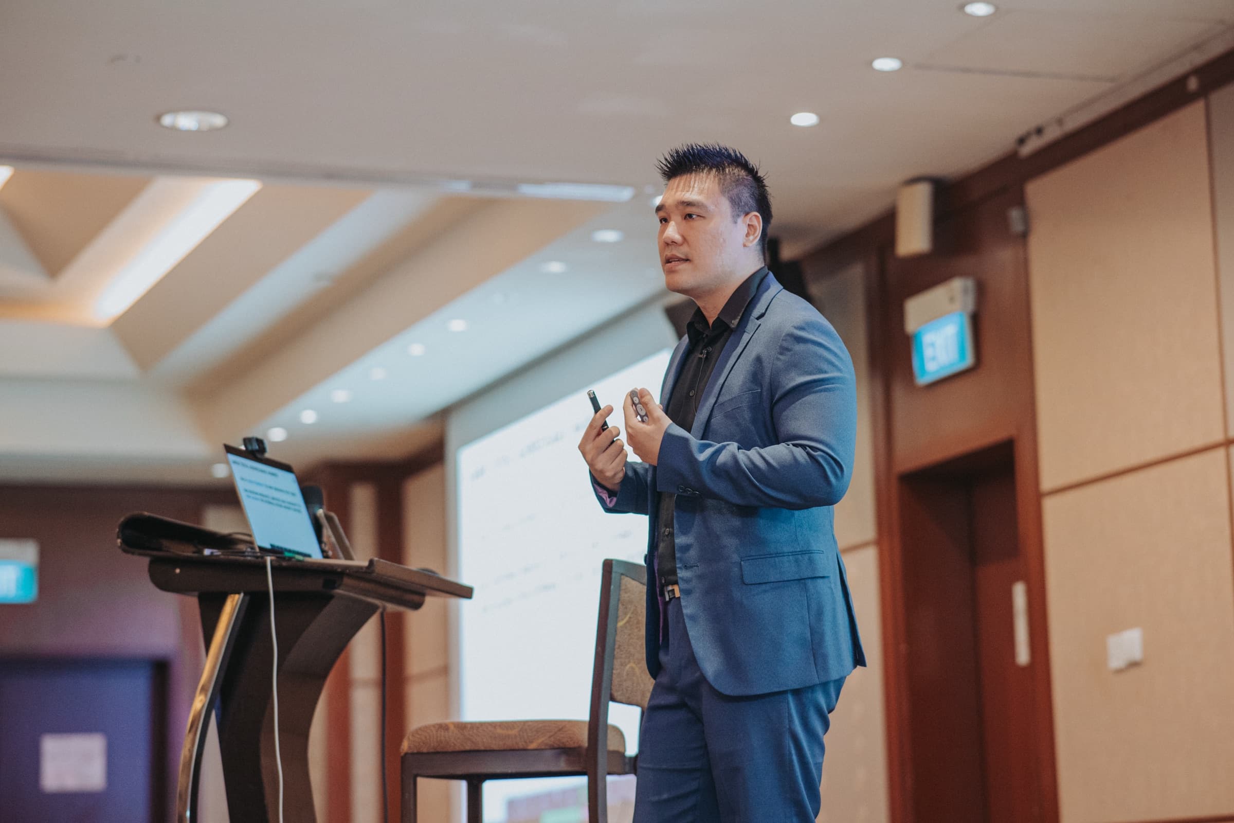 Daniel Lim in a navy suit presenting to investors with a slide clicker in hand during a live training session